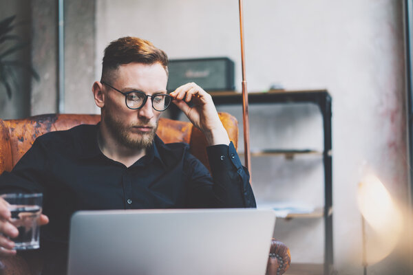 Бородатый молодой бизнесмен, работающий в современном лофт-офисе. Мужчина сидит в винтажном кресле, держа стеклянную воду. Использование современных ноутбуков, размытый фон. Горизонтальный, эффект пленки
.