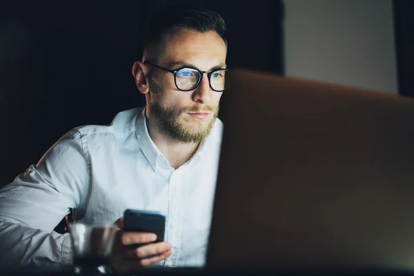 Retrato de hombre de negocios barbudo trabajando en la moderna oficina loft por la noche. Hombre usando teléfono inteligente contemporáneo, fondo borroso. Horizontal, efecto de película . —  Fotos de Stock