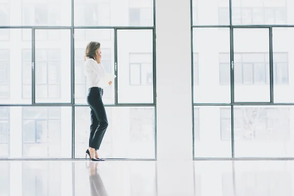 Foto mujer de negocios con traje moderno, teléfono inteligente hablando y sosteniendo papeles en las manos. Oficina de loft de espacio abierto. Fondo de ventanas panorámicas. Maqueta horizontal. . —  Fotos de Stock