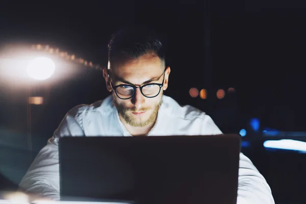 Empresario barbudo con gafas trabajando en la moderna oficina loft por la noche. Hombre utilizando el documento de texto portátil contemporáneo, fondo borroso. Horizontal, efecto película, bokeh —  Fotos de Stock