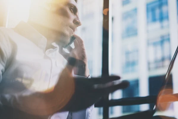 Foto closeup berjenggot pengusaha dewasa bekerja di kantor apartemen modern. Pria mengenakan kemeja putih dan menggunakan smartphone kontemporer. Latar belakang jendela panorama. Horisontal, efek film, bokeh — Stok Foto