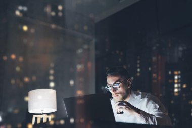 Geceleri modern çatı katında çalışan sakallı genç işadamı. Çağdaş not defteri mesajlaşma mesajını kullanan, elinde espresso, bulanık arka plan olan adam. Yatay, film efekti, bokeh