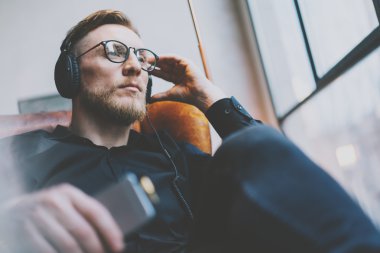 Portre yakışıklı sakallı adam gözlük, kulaklık müzik modern loft stüdyo dinlerken. Vintage sandalyede oturan, akıllı telefonu tutan ve dinlendirici bir adam. Panoramik pencereler arka plan. Yatay, film efekti