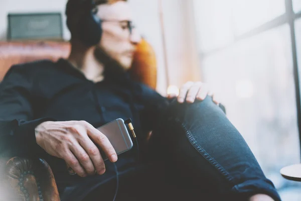 Foto bonito barbudo homem fones de ouvido ouvindo música moderna loft studio.Man sentado na cadeira vintage olhando janela, segurando smartphone e relaxing.Blurred fundo. Horizontal, efeito de filme . — Fotografia de Stock