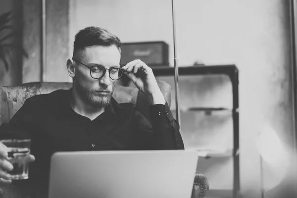 Giovane uomo d'affari barbuto che lavora in un moderno ufficio loft. Un uomo seduto su una sedia vintage, con in mano dell'acqua di vetro. Utilizzando notebook contemporaneo. Bianco e nero. Effetto orizzontale, pellicola . — Foto Stock