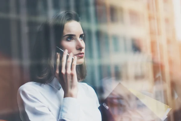 Imagen mujer de negocios con traje moderno, teléfono inteligente hablando y sosteniendo papeles en las manos. Oficina de loft de espacio abierto. Fondo de ventanas panorámicas. Una maqueta horizontal. Efecto película — Foto de Stock