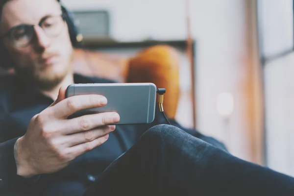 Close-up foto knap bebaarde man koptelefoon kijken video mobiele telefoon moderne loft Studio. Man zitten in Vintage stoel, het houden van smartphone en ontspannen. Horizontaal, filmeffect. — Stockfoto