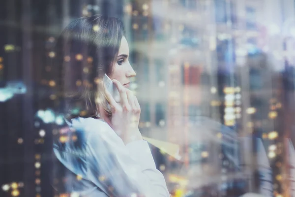 Foto wanita mengenakan kemeja putih, berbicara smartphone dan memegang rencana bisnis di tangan. Buka ruang kantor loteng. Jendela panorama, latar belakang kota malam. Mockup horisontal, bokeh. Efek film — Stok Foto