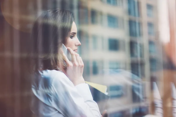 Foto mujer de negocios con traje moderno, teléfono inteligente hablando y sosteniendo papeles en las manos. Oficina de loft de espacio abierto. Fondo de ventanas panorámicas. Una maqueta horizontal. Efecto película —  Fotos de Stock