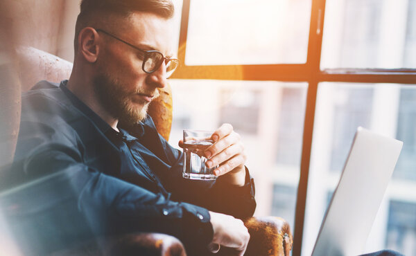 Крупный план фото бородатый бизнесмен, работающий на современном лофт-офисе. Мужчина сидит в винтажном кресле, держа стеклянную воду. Используя современный ноутбук, панорамный фон окон. Горизонтальный, эффект пленки
.