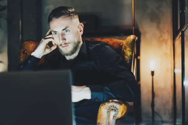 Foto hombre de negocios relajante oficina loft moderno. Hombre sentado en silla vintage por la noche. Usando contemporarylaptop, fondo borroso. Luz suave, sombras. Horizontal, efecto de película . —  Fotos de Stock