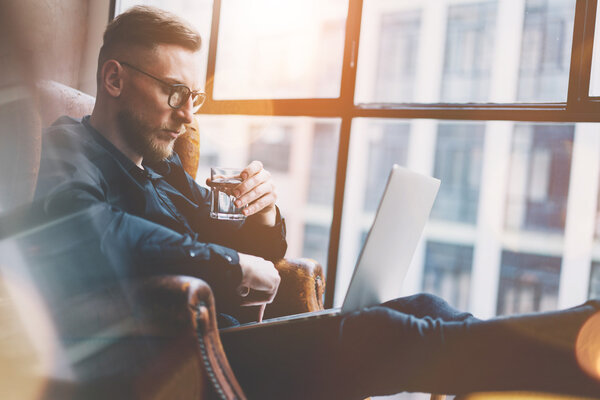 Фото бородатого бизнесмена, работающего в современном лофт-офисе. Мужчина сидит в винтажном кресле, держа стеклянную воду. Используя современный ноутбук, панорамный фон окон. Горизонтальный, эффект пленки
.