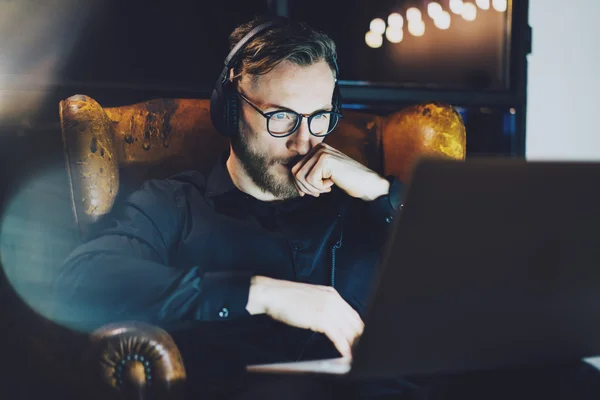 Foto barbudo hombre de negocios con gafas relajante moderno loft office.Man sentado en silla vintage, escuchar música portátil night.Using portátil contemporáneo, fondo borroso. Horizontal, efecto de película . —  Fotos de Stock