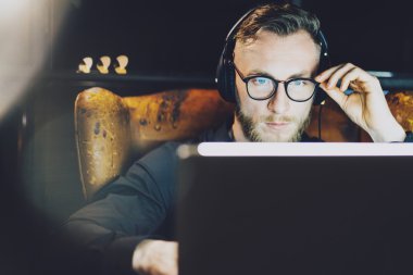 Çalışma modern loft office gözlüklü fotoğraf şık sakallı adam. Vintage sandalye, oturan genç bankacı dinleme müzik dizüstü gece. Çağdaş dizüstü kullanarak. Yatay, film etkisi