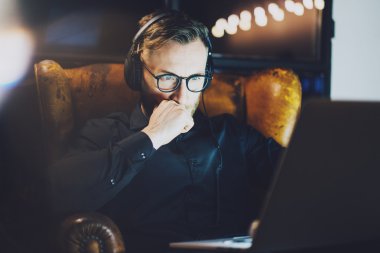 Sakallı adam gözlüklü çalışma modern loft office hayal et. Vintage sandalye, oturan genç bankacı dinleme müzik dizüstü gece. Çağdaş dizüstü bilgisayar kullanarak, arka plan bulanık. Yatay, film etkisi