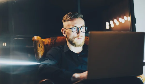 Picture bearded banker still working modern loft office after hard day.Man sitting in vintage chair at night.Using contemporary laptop, blurred background.Soft light,shadows.Horizontal,film effect — Stock Photo, Image