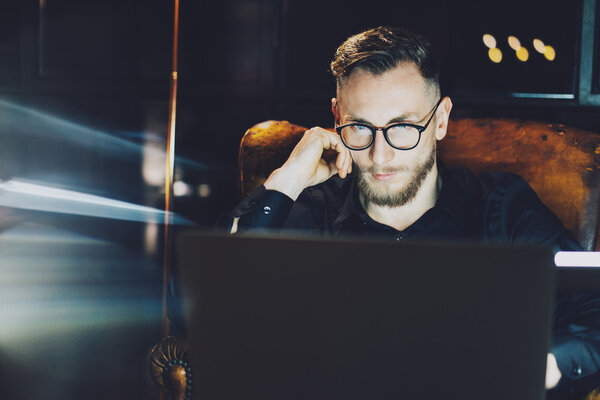 Молодой менеджер работает в современном лофт-офисе. Мужчина, сидящий ночью в винтажном кресле. Используя современный ноутбук, размытый фон. Мягкий свет, тени. Горизонтальный, эффект пленки
.