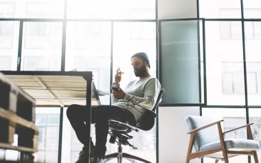 Fotoğraf sakallı adam yeni proje modern açık alan loft ile çalışıyor. Çağdaş cep telefonu ellerini kullanarak. Yaratıcı süreç. Panoramik pencere arka planı. Bulanık, film efekti, yatay
