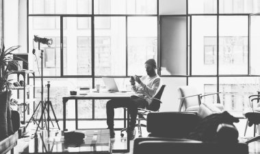 Fotoğraf sakallı yaratıcı yöneticisi yeni proje açık alan loft stüdyo ile çalışıyor. Modern akıllı telefon arıyorsunuz. Ahşap masa üzerinde Çağdaş dizüstü bilgisayar. Siyah ve beyaz. Bulanık arka plan, film efekti, yatay