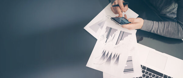 Working process. Account manager working at the table with new business project.Holding modern smartphone hand, statistics screen. Signs document and analyze plans. Wide,blurred, film effect