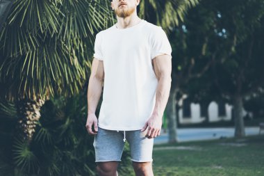 Fotoğraf Sakallı Kaslı Adam Yaz aylarında Beyaz Boş t-shirt ve şort giyiyor. Green City Garden Arka Plan, bulanık. Yatay Mockup