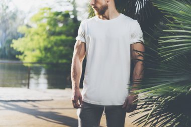 Fotoğraf Sakallı Kaslı Adam Yaz aylarında Beyaz Boş t-shirt giyiyor. Green City Garden, göl ve palmiye arka plan, bulanık. Yatay Mockup