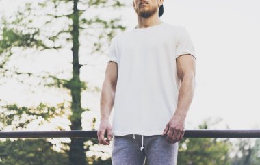 Fotoğraf Sakallı Kaslı Adam Beyaz Boş t-shirt, snapback kap ve şort yaz tatilinde giyiyor. Göl kenarında dinlendirici bir zaman. Green City Garden Park Sunset Arka Plan. Ön görüş. Yatay Mockup