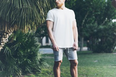 Fotoğraf Sakallı Kaslı Adam Yaz tatilinde Beyaz Boş t-shirt ve şort giyiyor. Sakin vakit. Green City Garden Park Arka Plan. Ön görüş. Yatay Mockup.