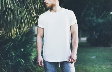 Fotoğraf Sakallı Kaslı Adam Yaz tatillerinde Beyaz Boş t-shirt ve şort giyiyor. Sakin vakit. Green City Garden Park Arka Plan. Ön görüş. Yatay Mockup.