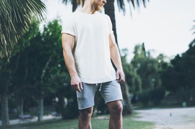 Görüntü Sakallı Kaslı Adam Yaz tatilinde Beyaz Boş t-shirt ve şort giyiyor. Sakin vakit. Green City Garden Park Arka Plan. Ön görüş. Yatay Mockup.