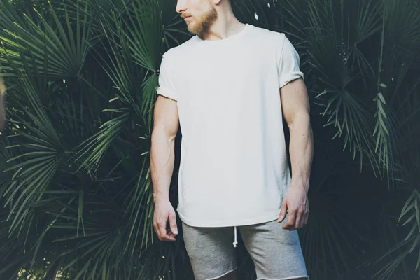 Photo Bearded Muscular Man Wearing White Blank t-shirt and shorts in summer time. Green huge palm Background. Horizontal Mockup — Stock Photo, Image