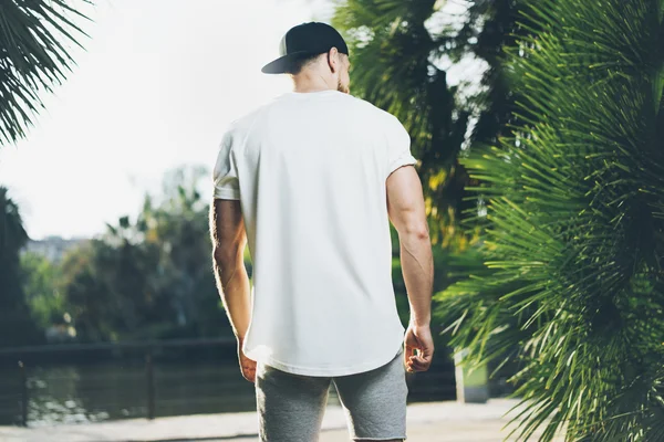 Zdjęcie Brodaty muskularny mężczyzna noszenie White blank t-shirt, czapka SnapBack i szorty w czasie letnim. Green City Garden Park tło. Widok z tyłu. Makieta pozioma — Zdjęcie stockowe