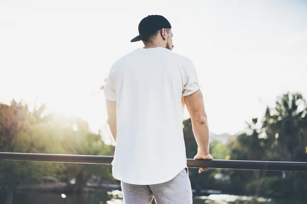 Photo Bearded Muscular Man T-shirt bianca con indosso bianco, berretto e pantaloncini in estate. Green City Garden Park Tramonto sfondo. Vista posteriore. Mockup orizzontale — Foto Stock