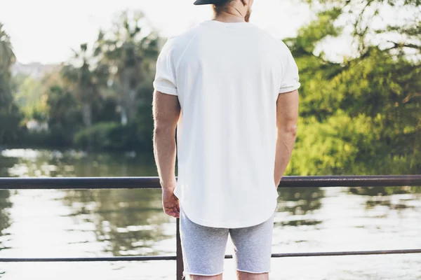 Foto Bearded gespierde man dragen wit blanco t-shirt, SnapBack Cap en shorts in de zomertijd. Ontspannen tijd in de buurt van het meer. Green City Garden Park zonsondergang achtergrond. Achteraanzicht. Horizontale mockup — Stockfoto