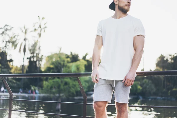 Bild skäggiga muskulös man bär vit blank t-shirt, snapback mössa och shorts i sommarsemester. Avkopplande tid nära sjön. Green City Garden Park Sunset bakgrund. tillbaka vy. Horisontellt utkast — Stockfoto