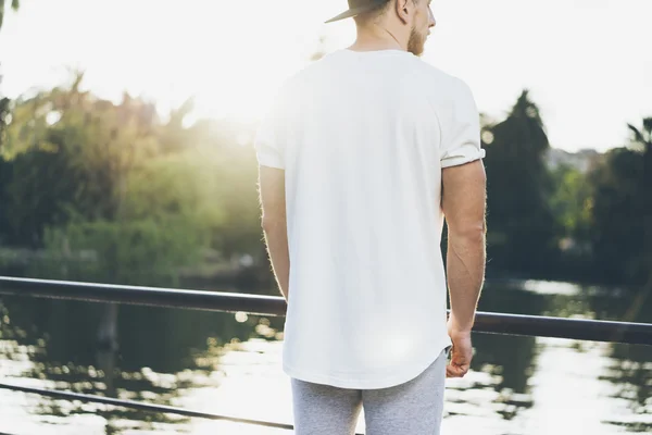 Picture Bearded Muscular Man Wearing White Blank t-shirt, snapback cap and shorts in summer vacation. Relaxing time near the lake. Green City Garden Park Sunset Background.Back view.Horizontal Mockup — Stock Photo, Image