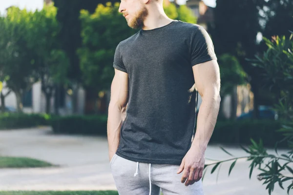 Foto Bearded muskulös man bär svart tom t-shirt och shorts i sommarsemester. Green City Garden Park bakgrund. Vy framifrån. Horisontellt utkast. — Stockfoto