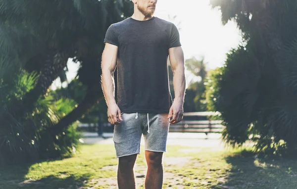 Fotografie Bărbat cu barbă musculară purtând tricou negru gol și pantaloni scurți în vacanța de vară. Walking Green City Garden Park Background. Privire din faţă. Mockup orizontal . — Fotografie, imagine de stoc