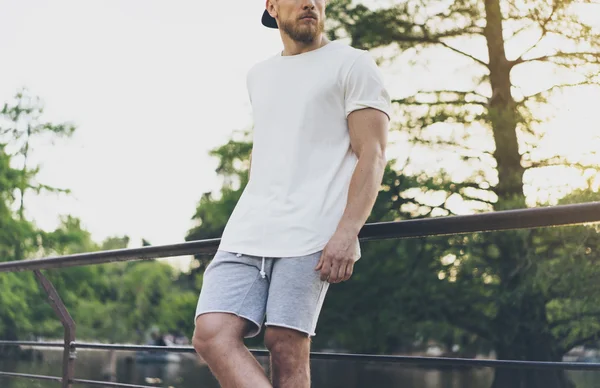 사진 수염 근육 남자 흰색 빈 티셔츠, 스냅백 모자와 여름 휴가반바지를 입고. 호수 근처에서 편안한 시간을 보내실 수 있습니다. 녹색 도시 정원 공원 일몰 배경입니다. 뒤로 보기. 수평 모형 — 스톡 사진