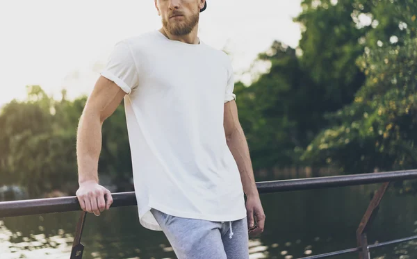 Foto Barbudo Muscular Hombre con blanco Camiseta vacía, gorra snapback y pantalones cortos en las vacaciones de verano. Tiempo relajante cerca del lago. Green City Garden Park Sunset Background (en inglés). Vista frontal. Mockup horizontal —  Fotos de Stock