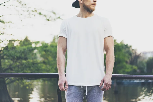 Foto Bearded muskulös man bär vit tom t-shirt, snapback mössa och shorts i sommarsemester. Kylning tid nära sjön. Green City Garden Park Sunset bakgrund. Vy framifrån. Horisontellt utkast. — Stockfoto