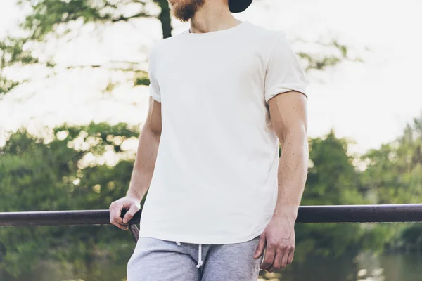 Kép szakállas izmos ember visel fehér üres póló, megtorló sapka és rövidnadrág nyári szünet. Hűtés idő közelében a tó. Green City Garden Park Sunset háttér. Elülső nézet. Vízszintes mockup. — Stock Fotó