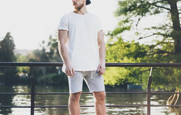 Kép szakállas izmos ember viselése fehér üres póló, megtorló sapka és rövidnadrág nyaralni. Hűtés idő közelében a tó. Green City Garden Park Sunset háttér. Elülső nézet. Vízszintes mockup. — Stock Fotó