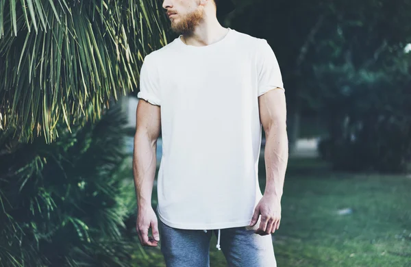 Foto Bearded muskulös man bär vit tom t-shirt och shorts i sommarsemester. Kylning tid. Green City Garden Park bakgrund. Vy framifrån. Horisontellt utkast. — Stockfoto