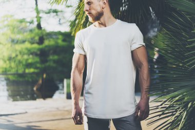 Fotoğraf Sakallı Kaslı Adam Yaz aylarında Beyaz Boş t-shirt giyiyor. Green City Garden, göl ve palmiye arka plan, bulanık. Yatay Mockup.