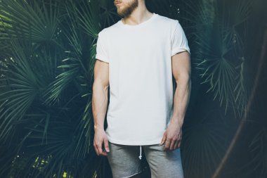 Fotoğraf Sakallı Kaslı Adam Yaz aylarında Beyaz Boş t-shirt ve şort giyiyor. Yeşil Ağaç Palmiye Arka Plan. Yatay Mockup.