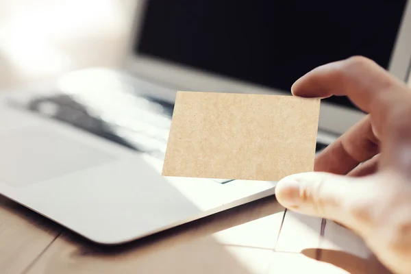 Close-up foto man toont lege ambachtelijke visitekaartje en met behulp van moderne laptop houten tafel. Onscherpe achtergrond. Mockup klaar voor Priva Infortion. Zonlicht reflecties oppervlak gadget. Horizontale mock up. — Stockfoto