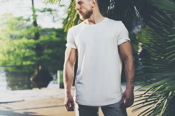 Fotografický vousatý svalnatý muž v letním čase nosí bílé tričko. Zelená městská zahrada, jezero a palmy na pozadí, rozmazané. Horizontální mockup. — Stock fotografie