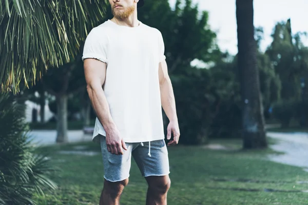 Foto Homem Muscular Barbudo Vestindo t-shirt branca em branco e shorts no verão. Green Tree Palm, fundo desfocado. Mockup horizontal . — Fotografia de Stock