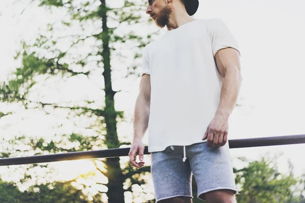 Immagine Uomo muscoloso barbuto Vestito con t-shirt bianca, berretto e pantaloncini in vacanza estiva. Green City Garden Park Sfondo.Vista anteriore.Mockup orizzontale . — Foto Stock